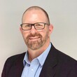 A headshot of a white, middle aged man wearing a suit, glasses, and smiling.