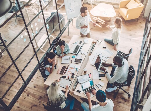 Group of employees gathered around a table in a bright office surrounded by smart glass windows.